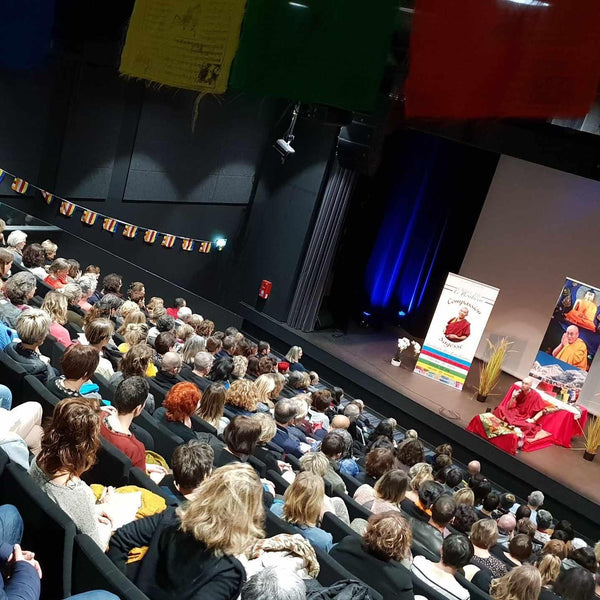 Conférence sur la méditation bouddhiste avec un lama sur scène devant un public, drapeaux tibétains et affiches de compassion
