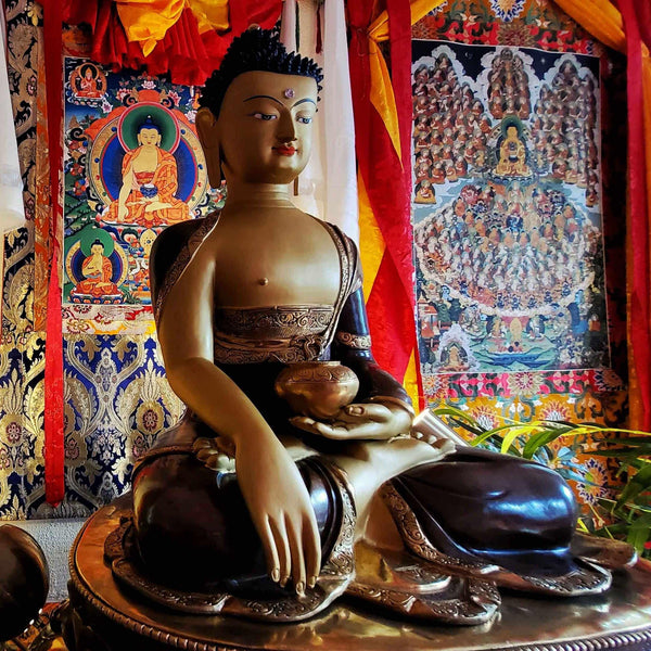 Statue bouddhiste tibétaine assise avec bols, devant thangkas colorés et tissus rituels