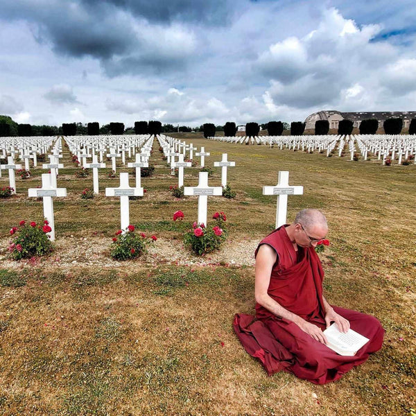 Moine bouddhiste en robe rouge méditant parmi des croix blanches dans un cimetière sous un ciel nuageux