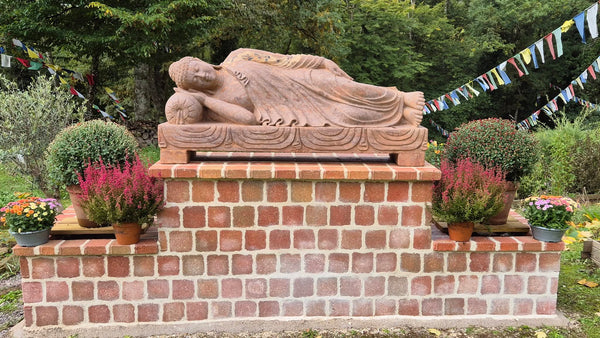 Statue bouddhiste couchée en pierre, drapeaux tibétains et fleurs dans un jardin paisible
