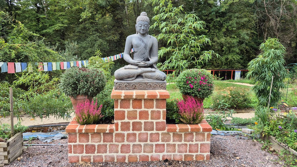 Statue de Bouddha en méditation sur socle en briques, jardin zen avec drapeaux tibétains et fleurs