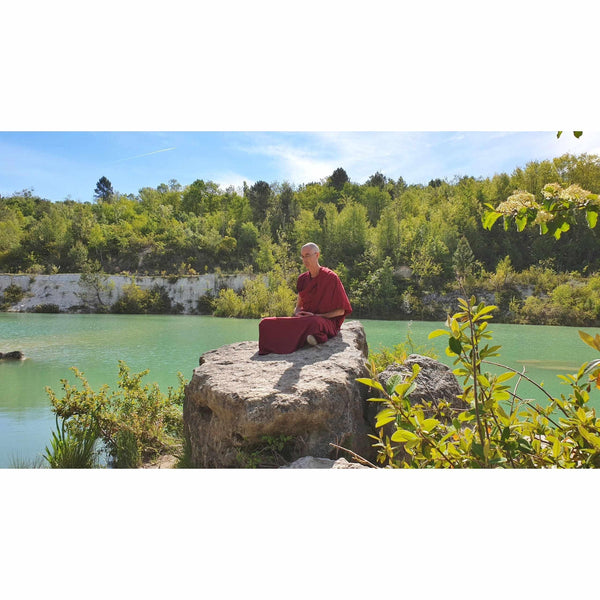 Moine bouddhiste en méditation sur rocher au bord d'un lac, vêtu de robe rouge, forêt en arrière-plan