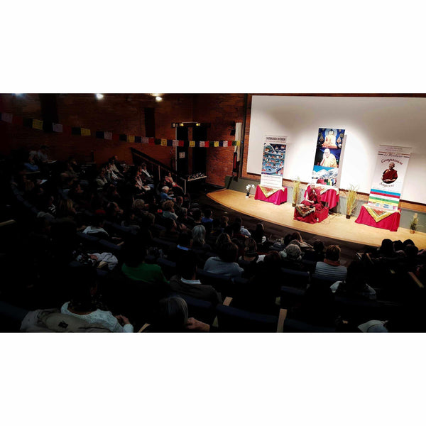 Conférence de méditation bouddhiste avec enseignant sur scène, public attentif, drapeaux tibétains