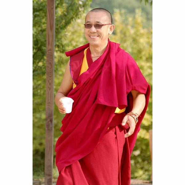 Lama bouddhiste en robe rouge souriant, tenant une tasse, dans un cadre naturel verdoyant