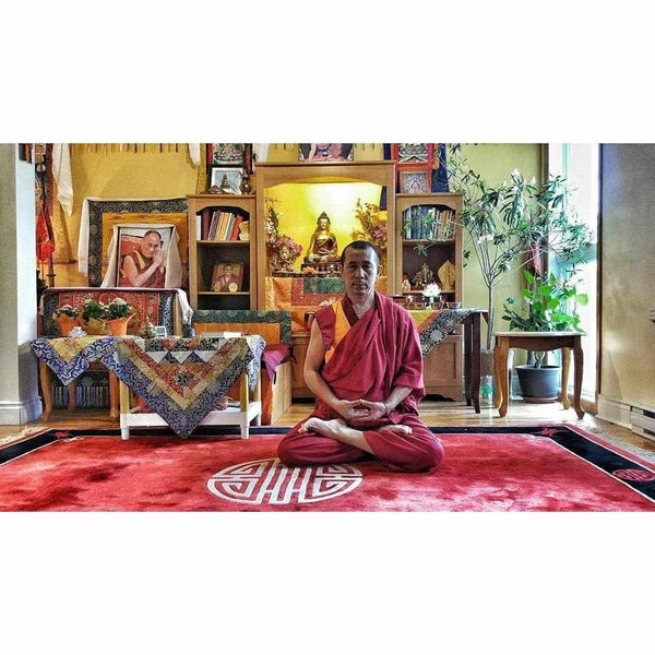 Moine bouddhiste en méditation sur tapis rouge, statue de Bouddha et objets tibétains en arrière-plan