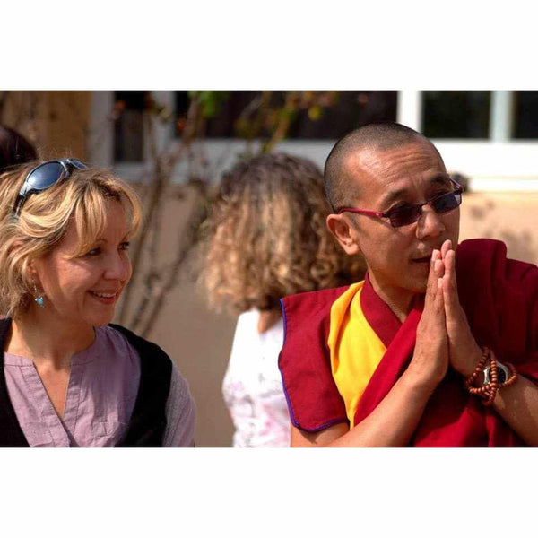 Lama bouddhiste en robe rouge et jaune, mains jointes en prière, avec une femme souriante en extérieur.