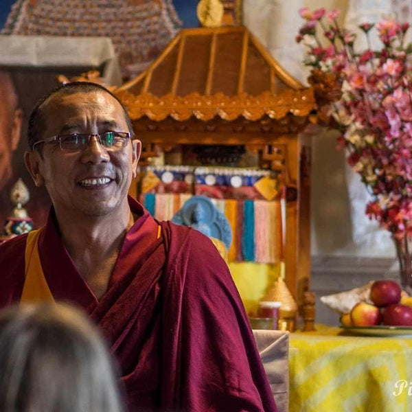 Lama bouddhiste souriant en robe rouge, devant autel tibétain avec offrandes et fleurs, enseignant la méditation
