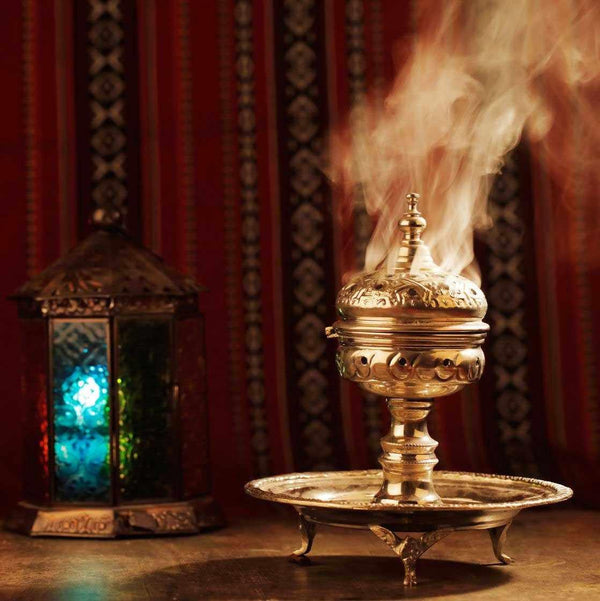 Brûle-encens bouddhiste tibétain en métal doré avec fumée, devant rideau rouge et lanterne colorée