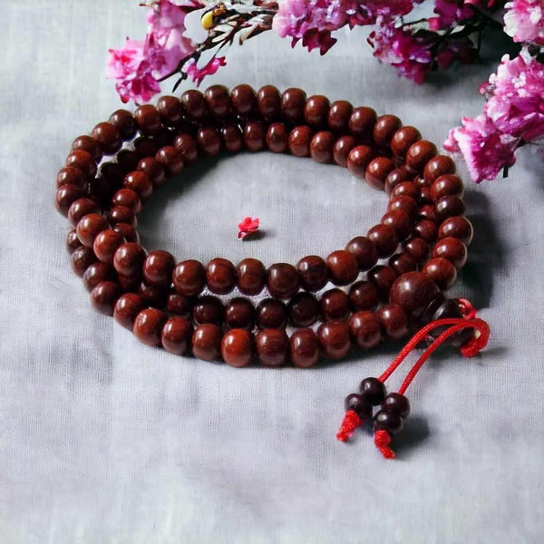 Chapelet bouddhiste en bois rouge avec nœuds rouges, fleurs roses en arrière-plan, symbole de méditation tibétaine