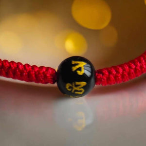 Bracelet tibétain rouge avec perle noire gravée de symboles bouddhistes dorés, objet de méditation et spiritualité.