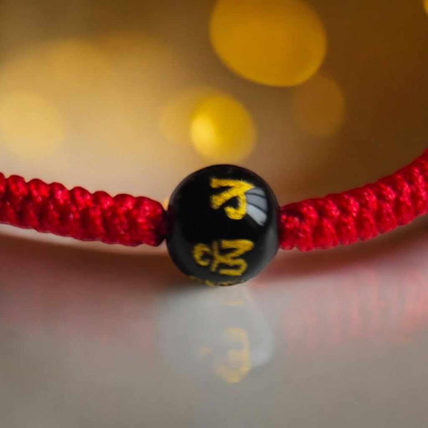 Bracelet tibétain rouge avec perle noire gravée de symboles bouddhistes dorés, objet de méditation et spiritualité.
