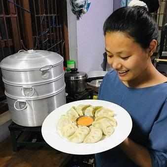 Femme souriante tenant un plat de momos tibétains frais, accompagnés d'une sauce orange, dans une cuisine traditionnelle.
